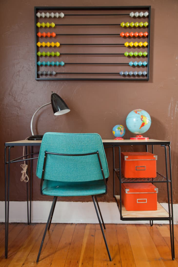 Teal chair at a desk with a black lamp, globes, orange boxes, and a colorful abacus on a brown wall.