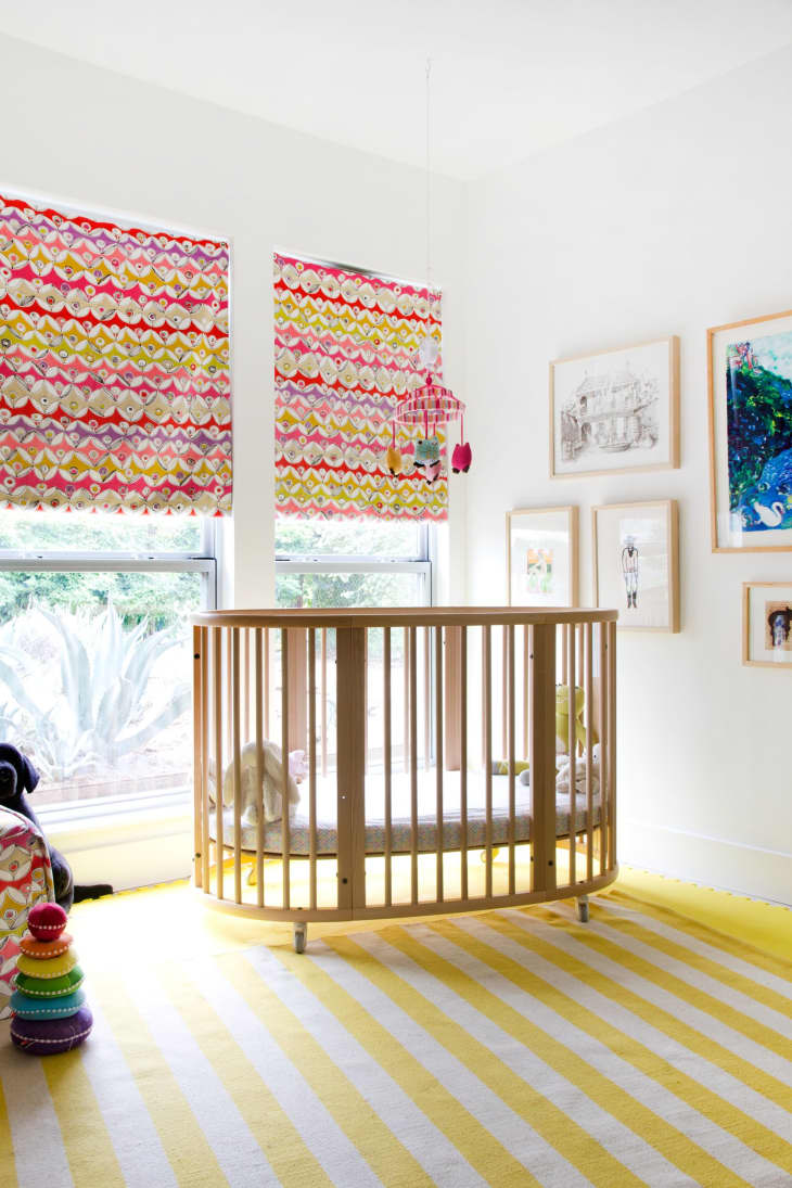 Nursery with a round wooden crib, colorful blinds, striped yellow rug, and wall art.