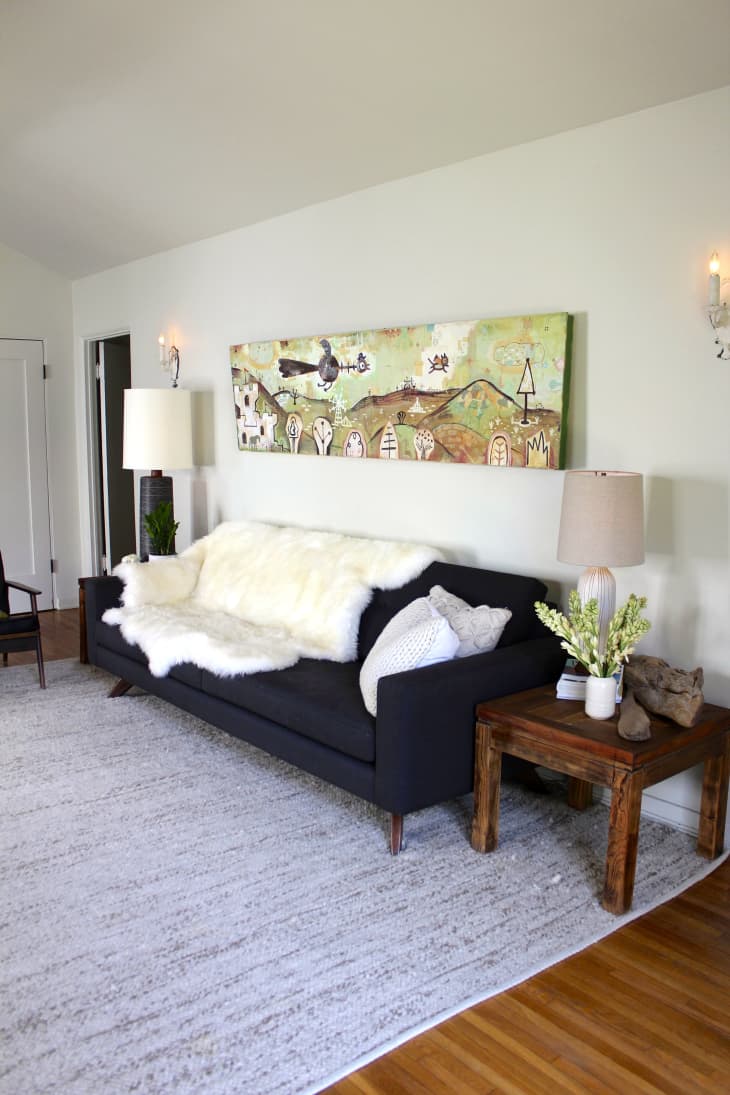 Black sofa with white fur throw, wooden side table, lamps, and abstract wall art in a living room.