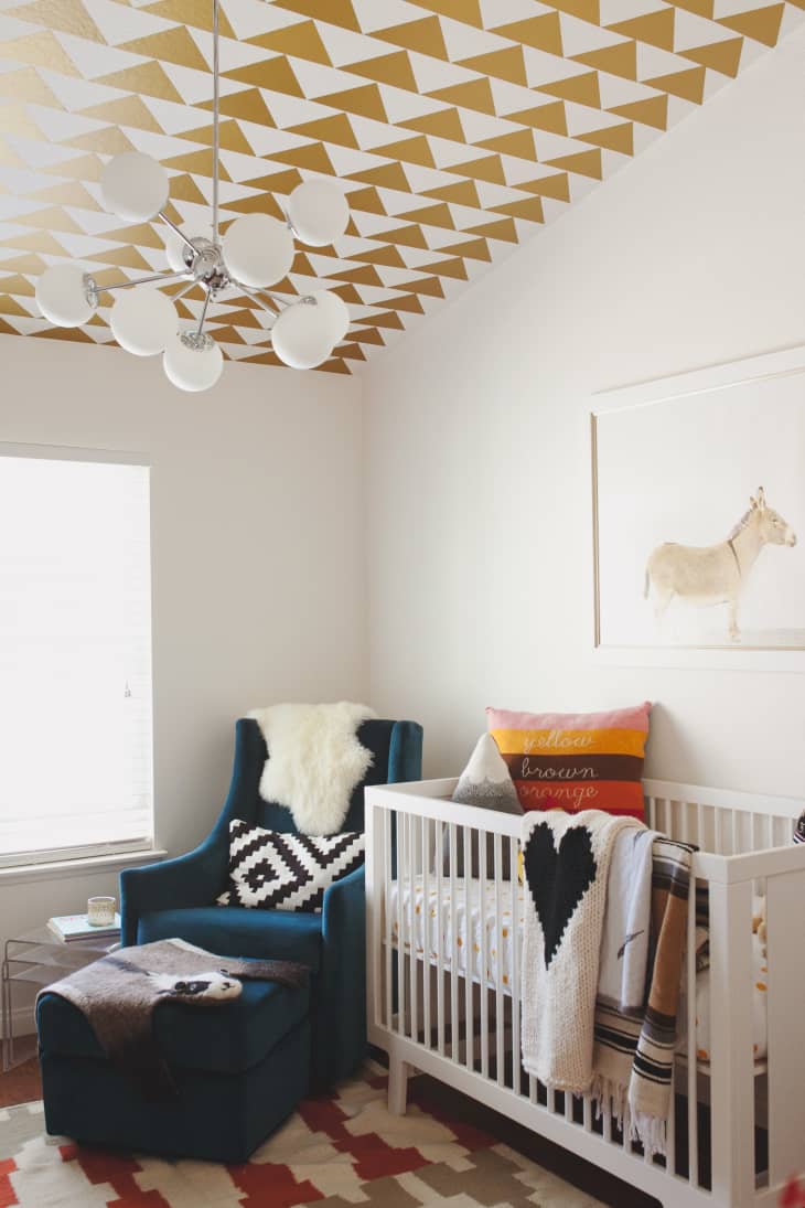 Nursery with geometric ceiling, blue armchair, white crib, and colorful pillows.