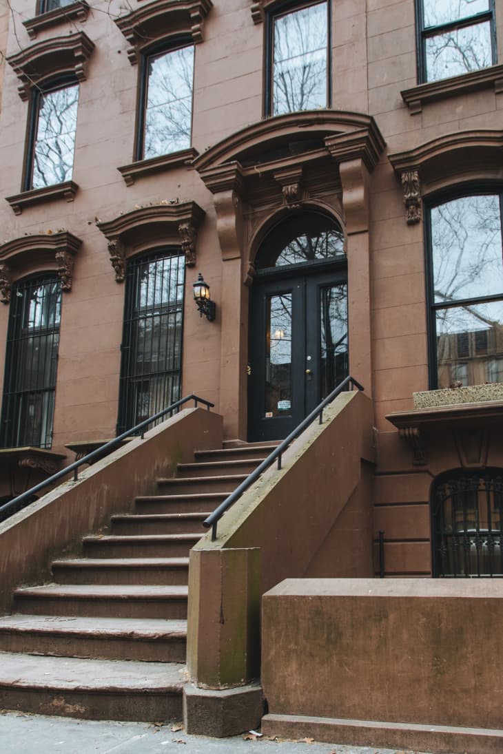 Brownstone building facade with arched doorway, black door, and steps leading up, flanked by wrought iron railings.