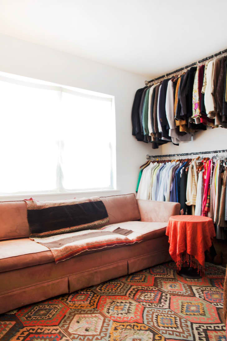 Pink sofa with patterned blankets, red tablecloth, and colorful clothes on racks in a bright room.