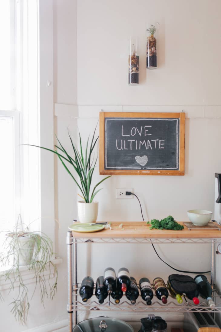 Kitchen corner with a chalkboard saying "Love Ultimate," potted plants, wine rack, and a wooden shelf with bowls and greens.