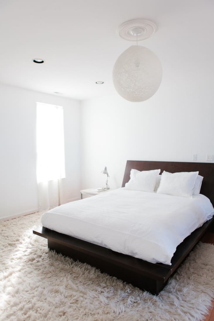 Minimalist bedroom with a dark wood bed frame, white bedding, fluffy rug, and a spherical ceiling light.