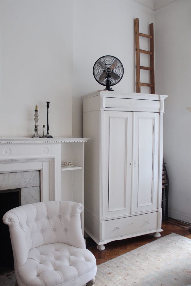 White armchair, vintage wardrobe with fan on top, fireplace with candlesticks, and wooden ladder in a cozy room.