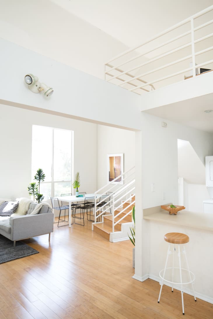 Open-plan living area with a gray sofa, dining table, white staircase, and bar stool on wooden flooring.