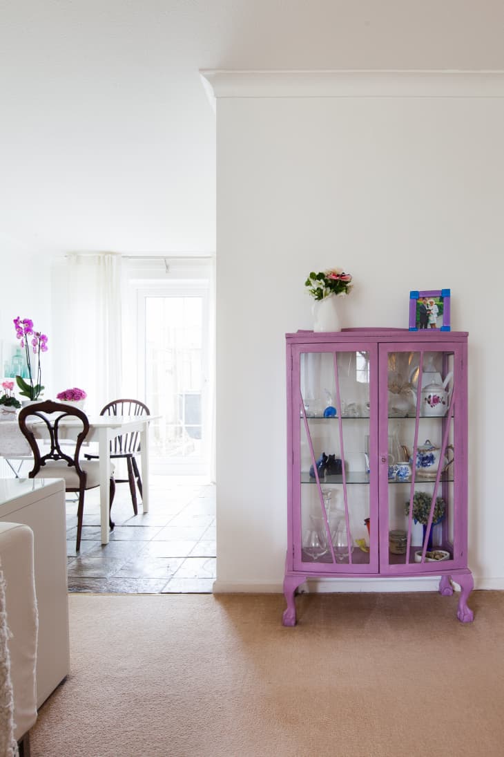 Purple display cabinet with glass doors, holding various decorative items, next to a dining area with wooden chairs.