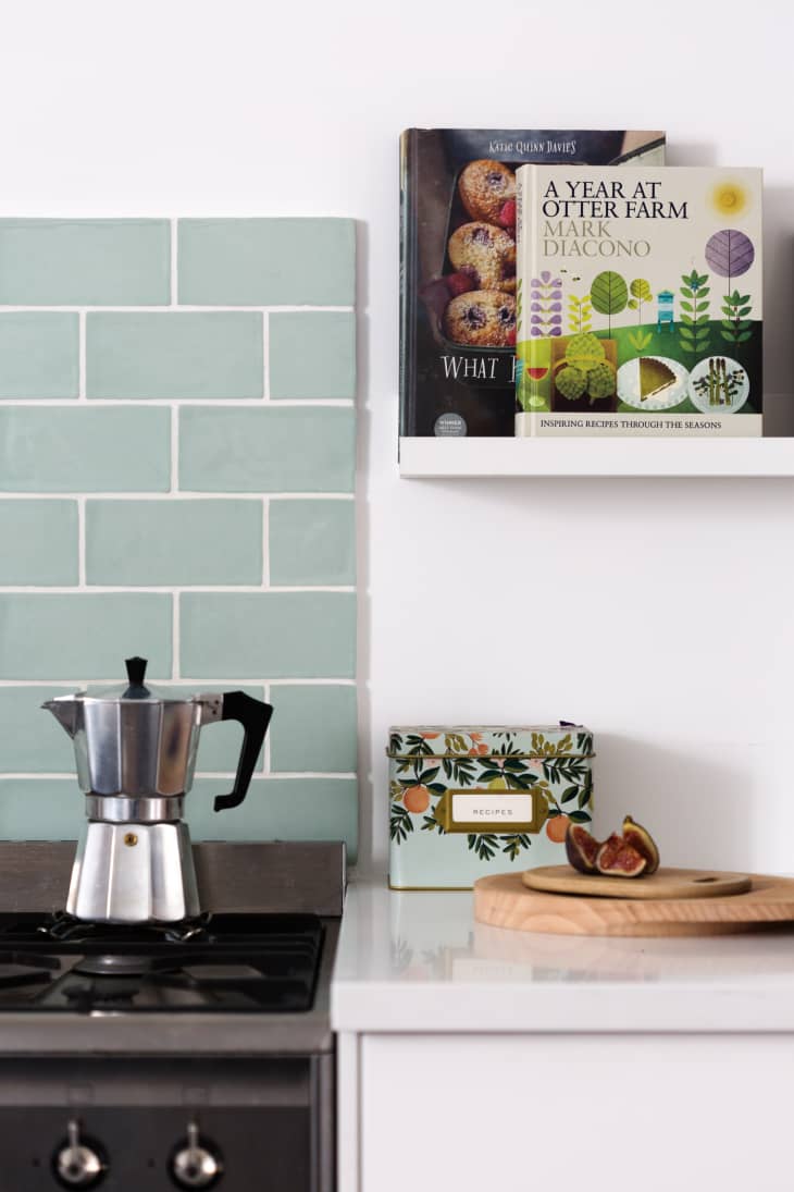 Stovetop espresso maker on kitchen counter with green tiles, recipe box, and sliced figs on a wooden board.