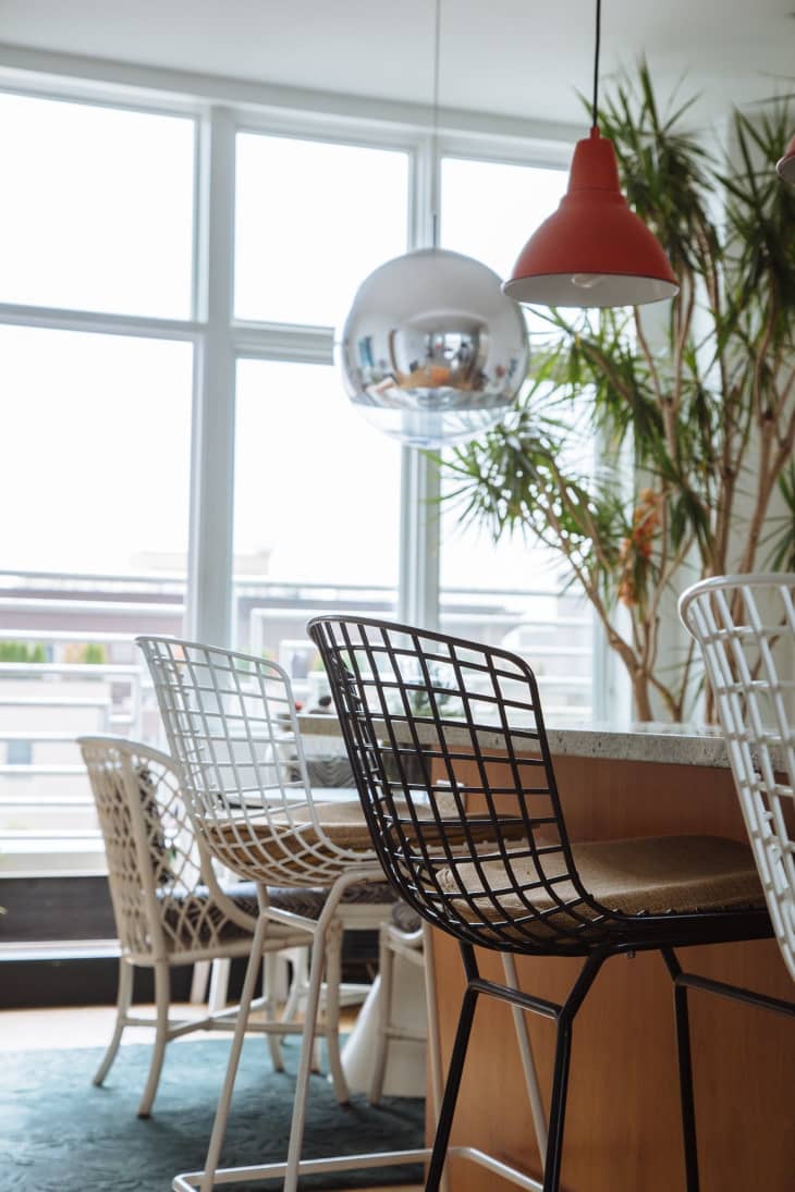 Modern kitchen with wire bar stools, marble countertop, red and silver pendant lights, and large potted plant.