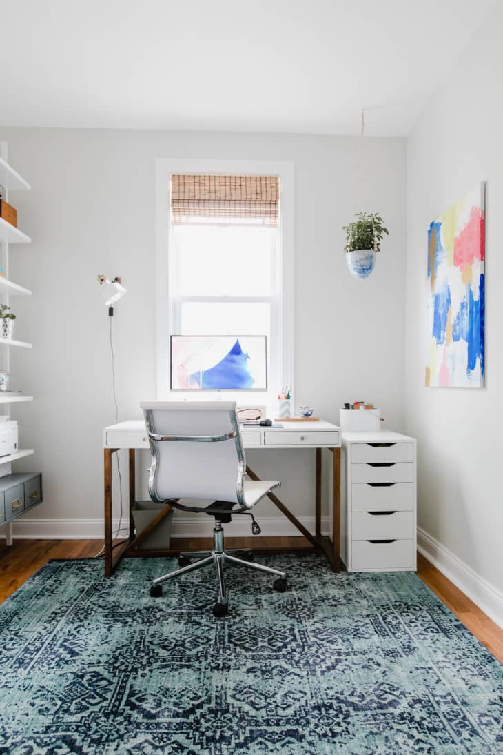 Home office with white desk, swivel chair, abstract art, and blue patterned rug.