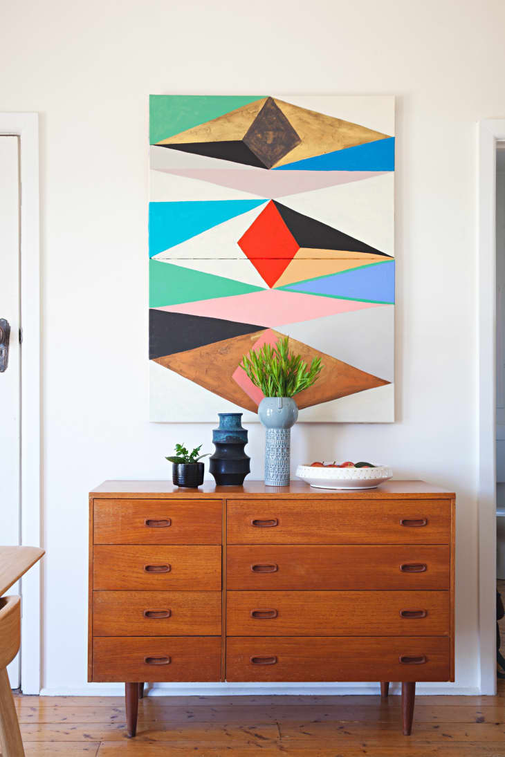 Mid-century wooden dresser with vases and a fruit bowl, beneath a colorful geometric wall art piece.