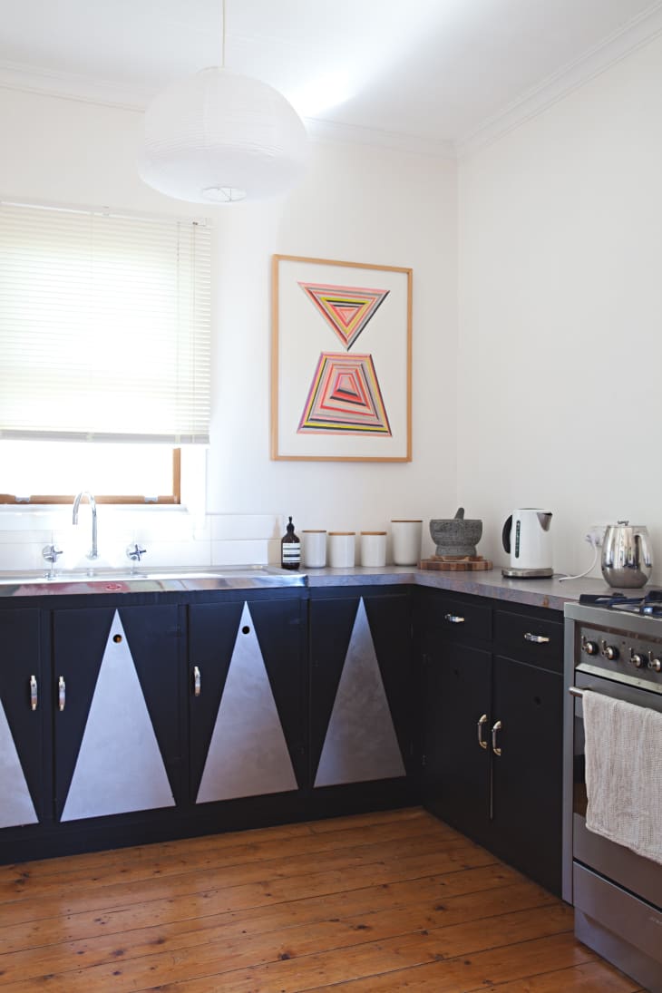 Modern kitchen with black cabinets featuring silver triangle designs, wooden floor, colorful geometric artwork, and stainless steel appliances.