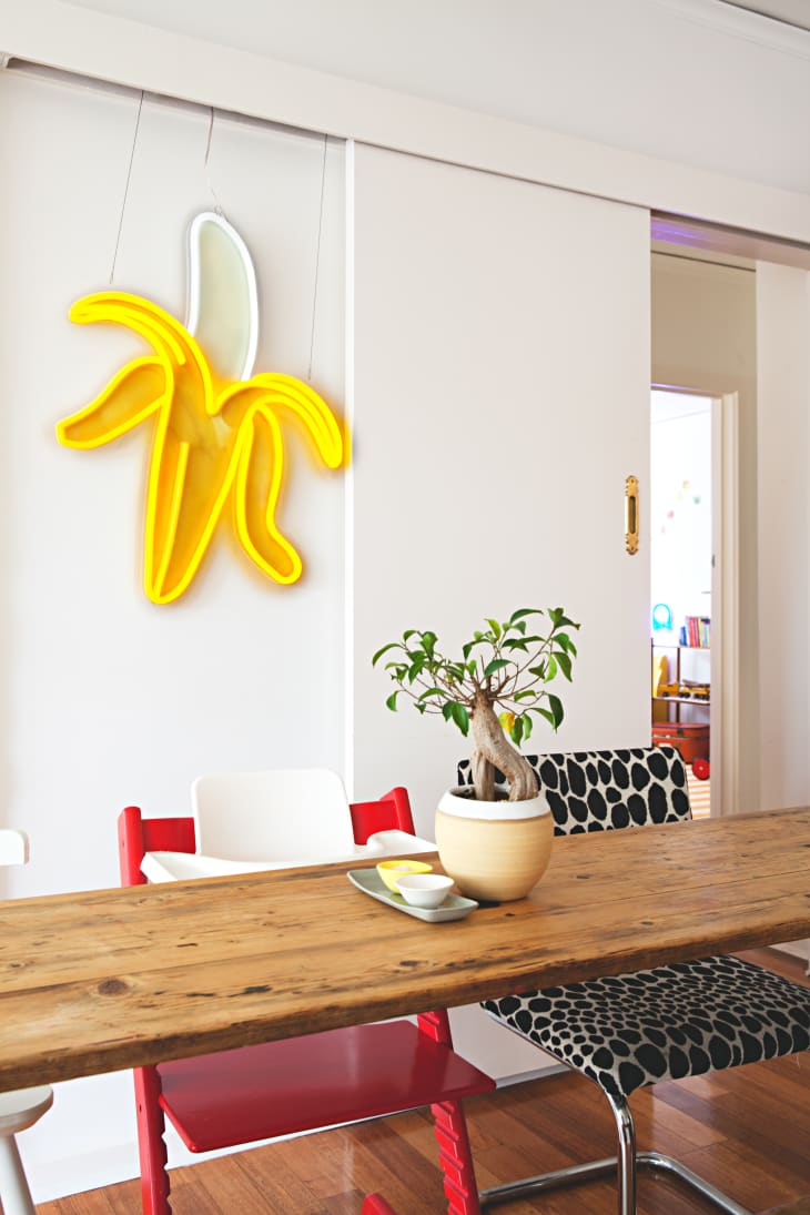 Dining area with wooden table, red and white chairs, potted plant, and yellow banana neon light on the wall.
