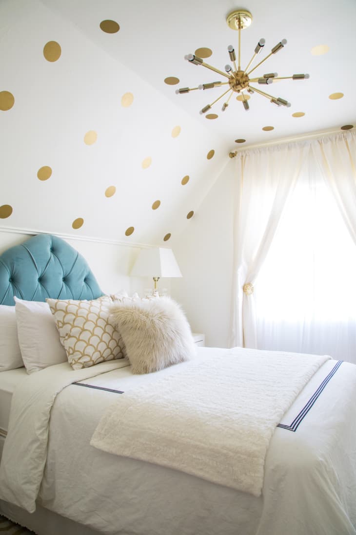 Bedroom with blue tufted headboard, gold polka dot wall, fluffy pillows, and modern chandelier.