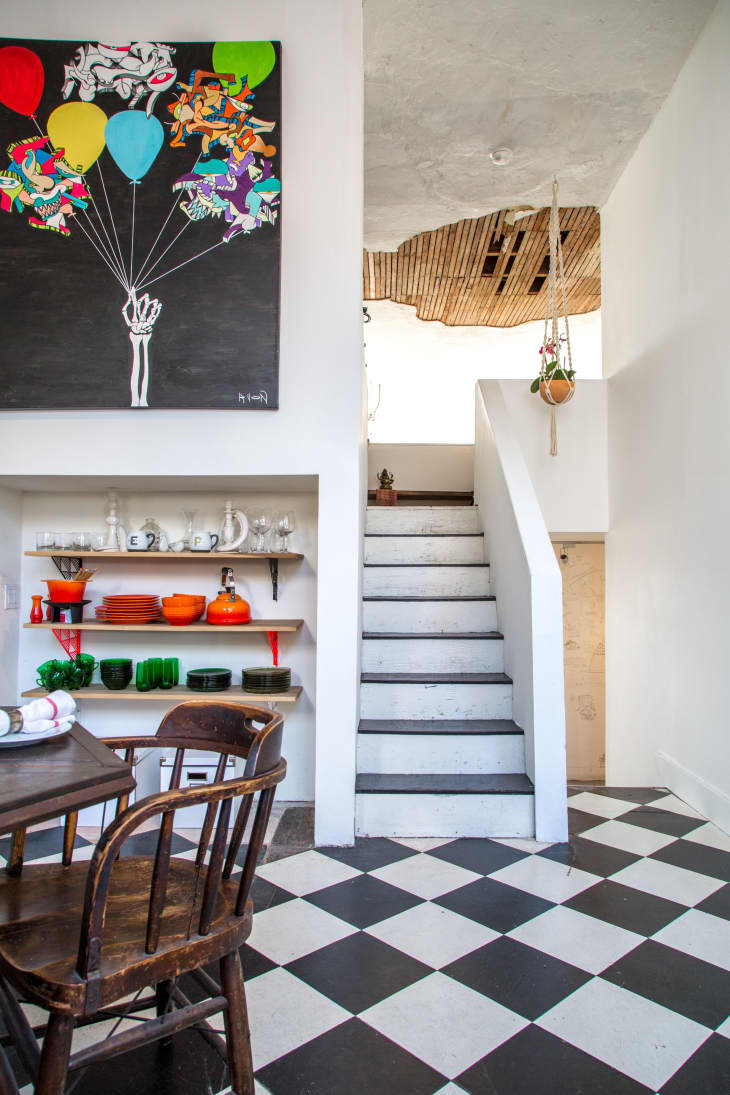 Dining area with checkered floor, colorful dishware on shelves, and a staircase leading to a loft with hanging plants.