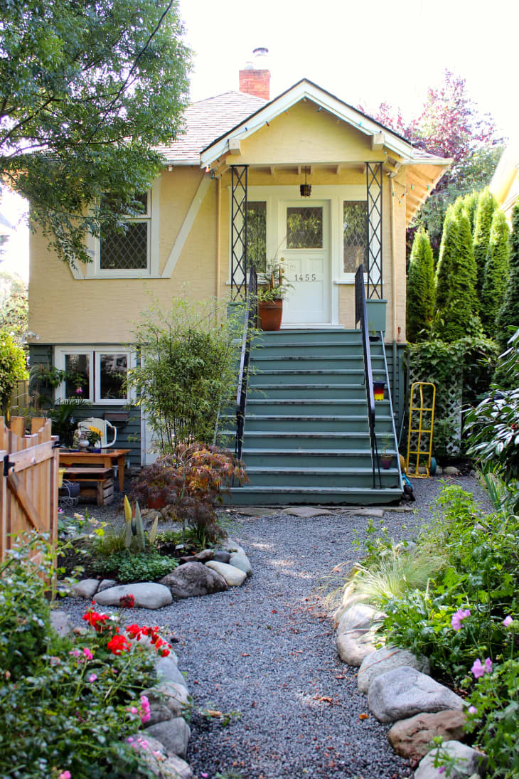 Yellow house with green steps, surrounded by lush garden, red flowers, and stone path leading to the entrance.
