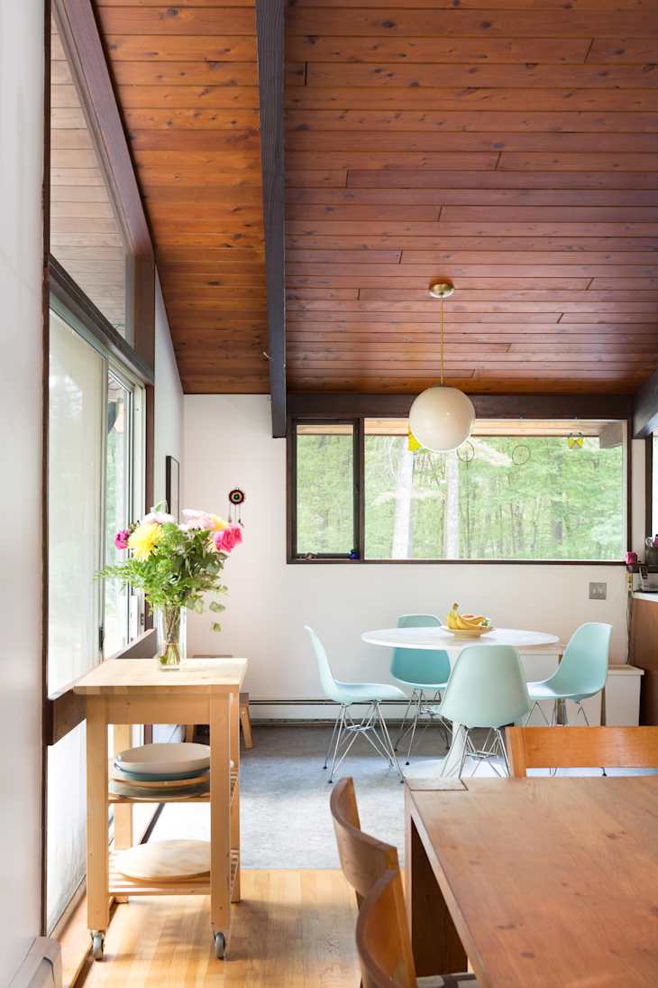 Mid-century modern dining room with wooden ceiling, round table, blue chairs, and a vase of flowers on a cart.