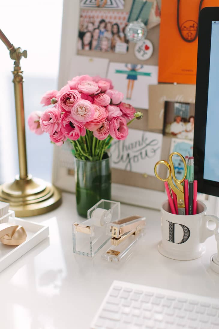 Pink ranunculus bouquet on a desk with a lamp, keyboard, and stationery in a mug.