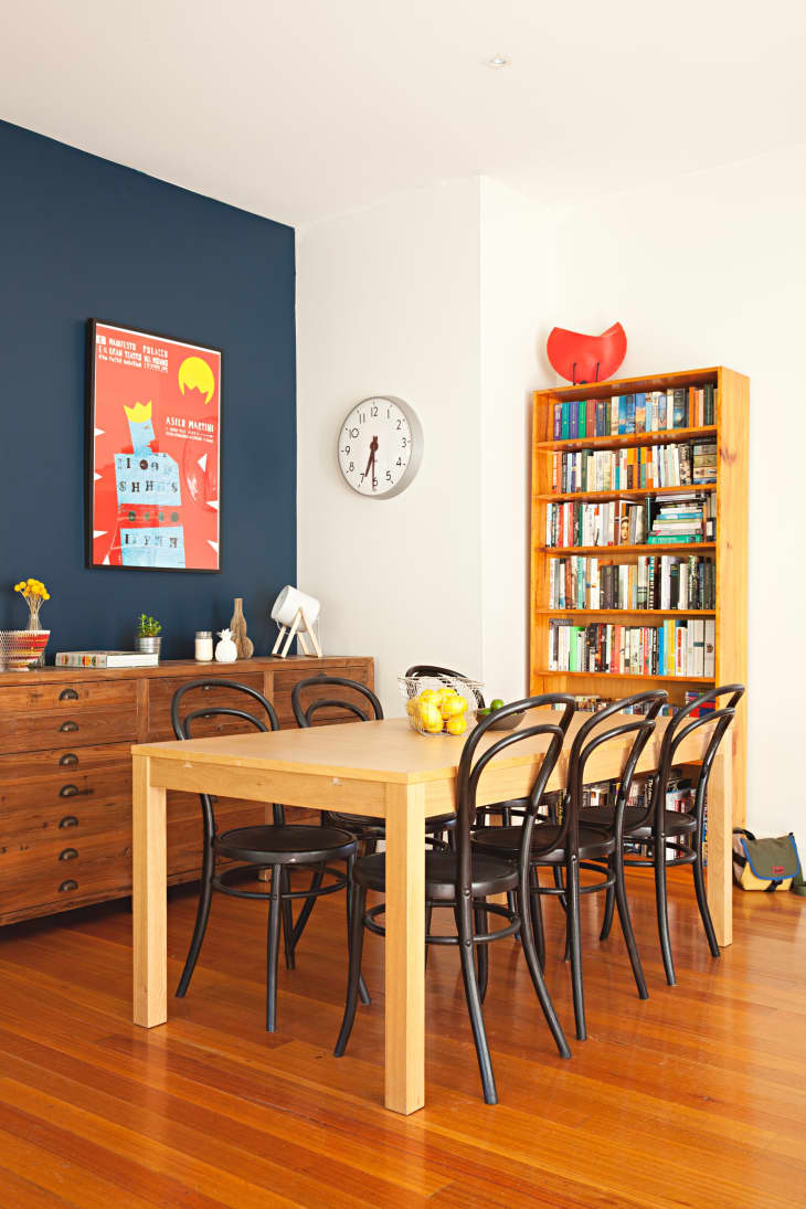 Dining room with wooden table, black chairs, bookshelf, wall clock, and colorful poster on a blue accent wall.