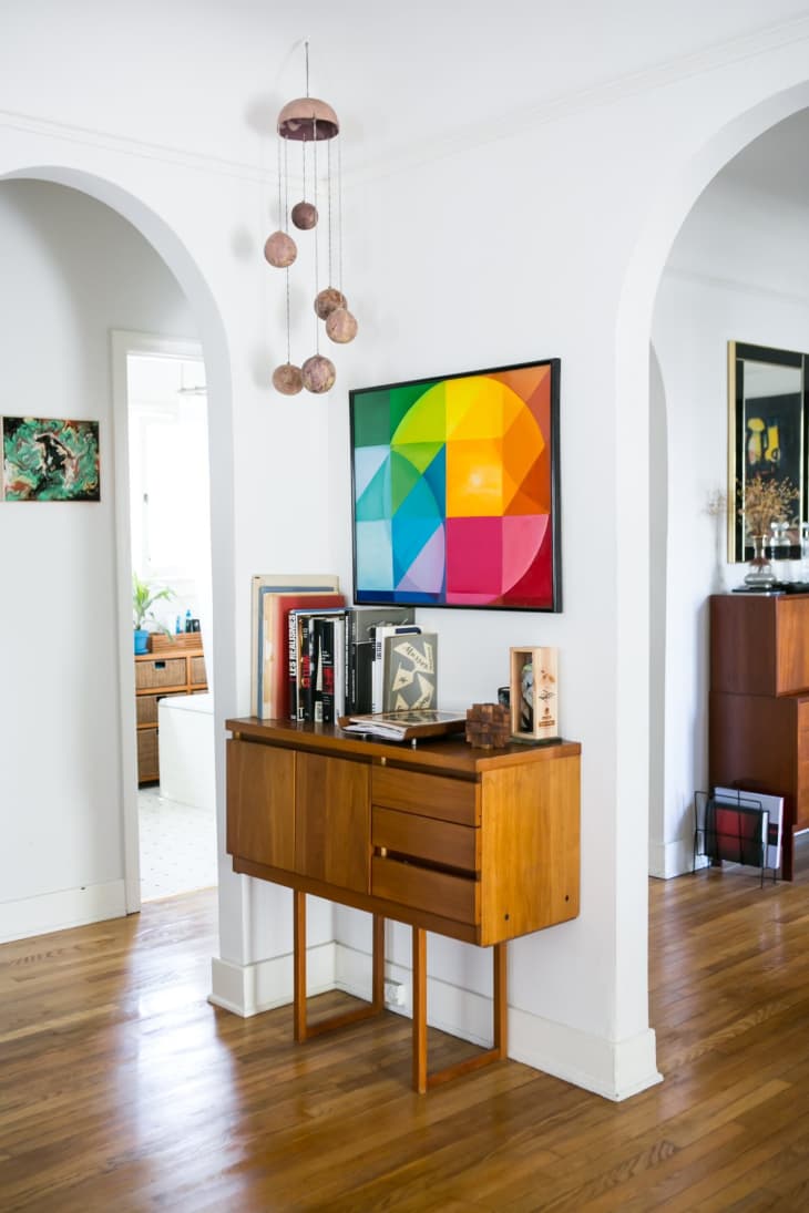 Mid-century modern console with books and decor, under a colorful geometric artwork, in a bright hallway.