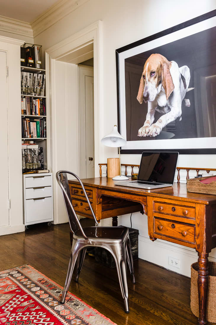 Home office with wooden desk, metal chair, laptop, desk lamp, and large dog portrait on the wall.