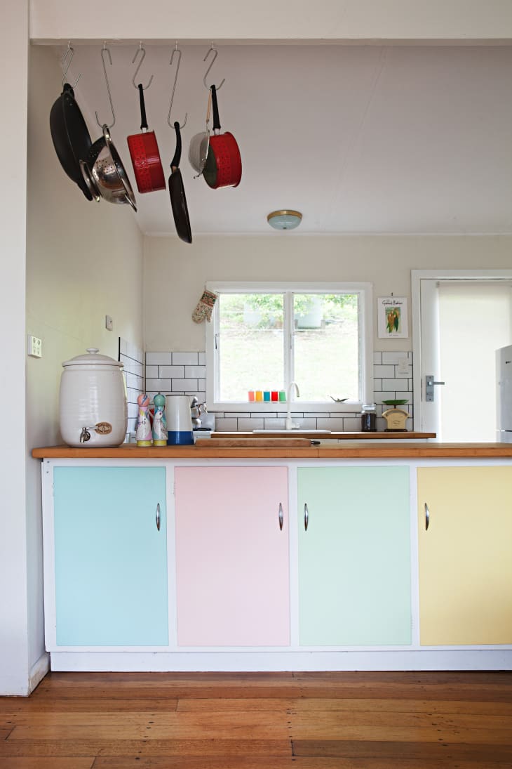 Pastel-colored kitchen cabinets with hanging pots, wooden countertop, and a window with a view of greenery.