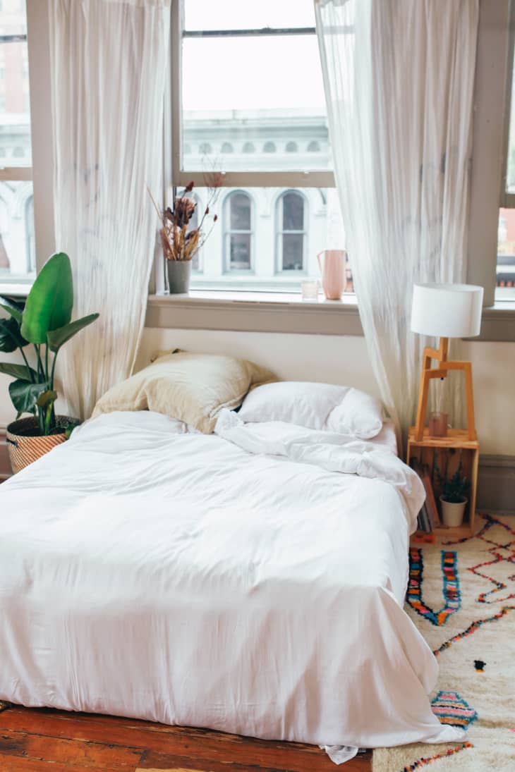 Cozy bedroom with unmade bed, white bedding, large windows, potted plant, and a wooden lamp on a colorful rug.