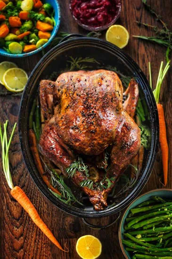 Roasted turkey in a black pan with herbs, surrounded by carrots, lemon halves, green beans, and a bowl of mixed vegetables.