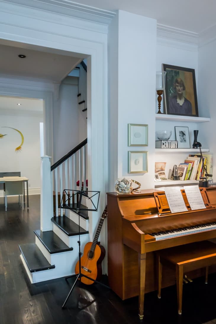 Wooden piano with sheet music, guitar, and music stand beside a staircase in a cozy living room.