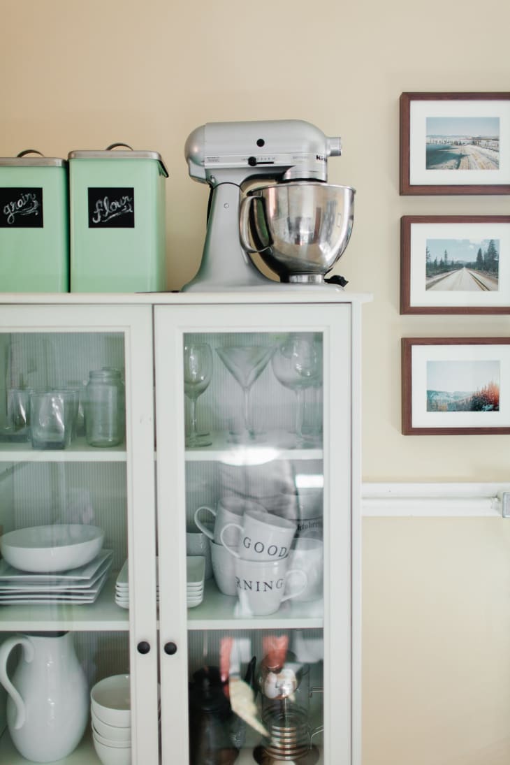 Kitchen cabinet with glass doors, mixer on top, green canisters, and framed photos on the wall.