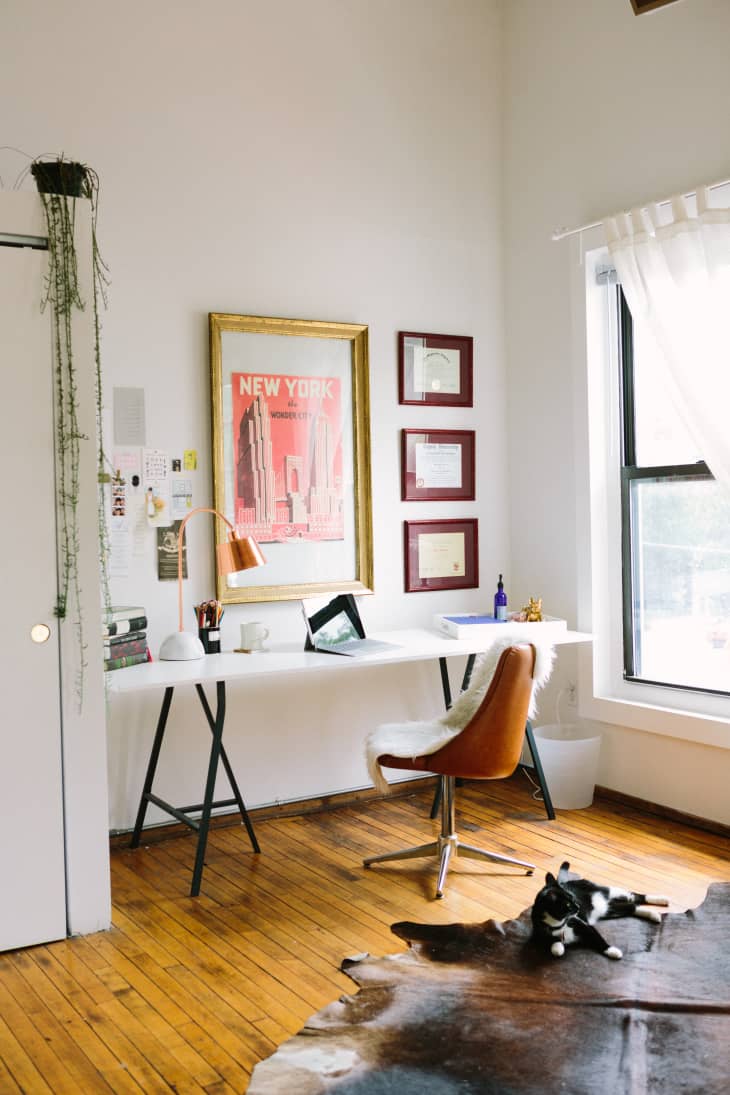 Home office with a white desk, brown chair, New York poster, certificates, and a cat on a cowhide rug.