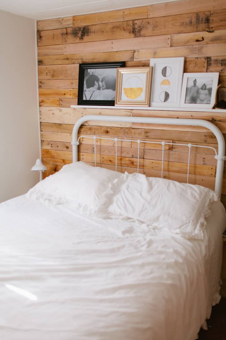 a bed with white bedding and wrought iron headboard sits against a pallet wood wall