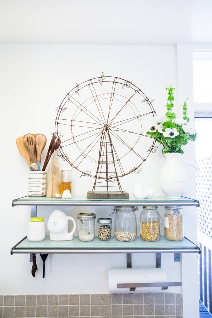 Kitchen shelves with a wire Ferris wheel, wooden utensils, jars of grains, and a white vase with green flowers.