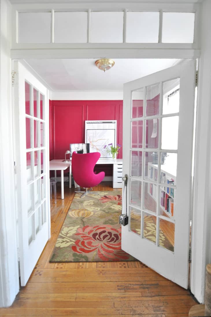French doors open to a home office with a pink accent wall, pink chair, floral rug, and white desk.
