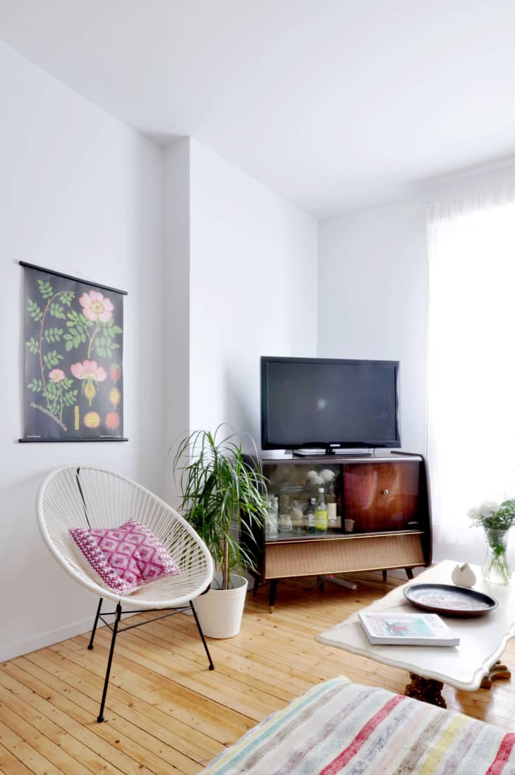 Living room with a TV on a vintage cabinet, white chair with pink cushion, potted plant, floral wall art, and striped rug.