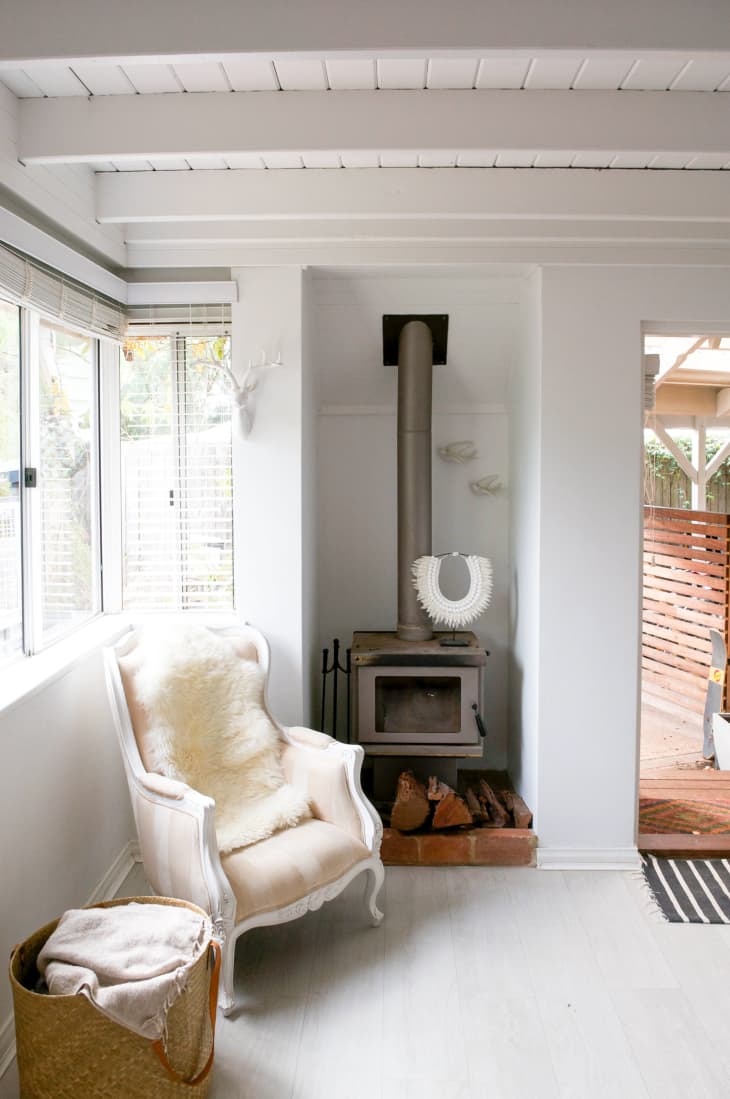 Cozy corner with a white armchair, sheepskin throw, wood stove, and basket in a bright room with large windows.