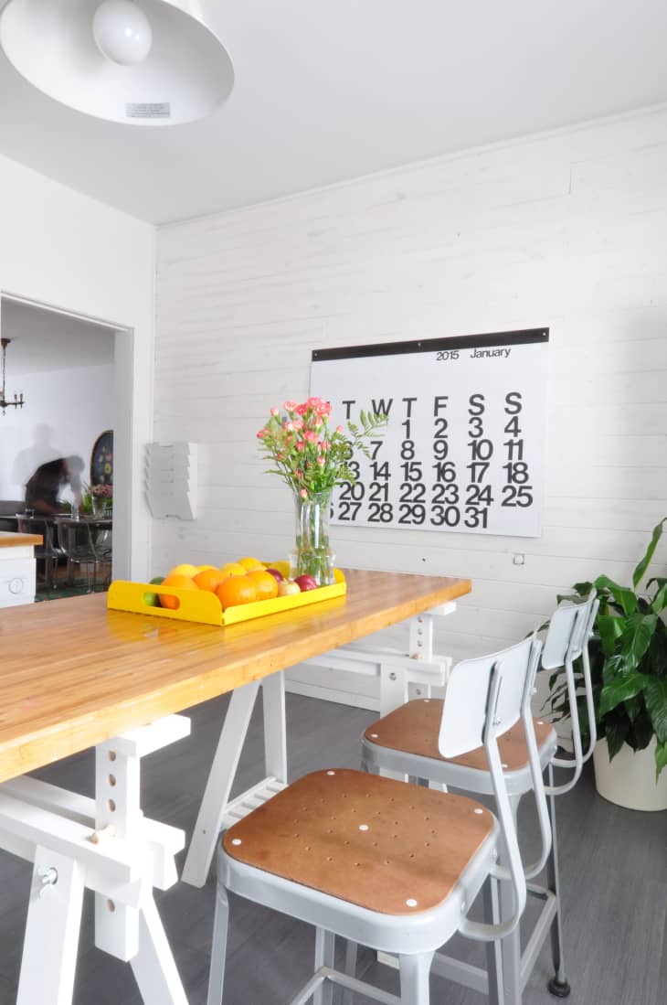 Dining area with wooden table, metal chairs, a vase of flowers, fruit tray, and large wall calendar.