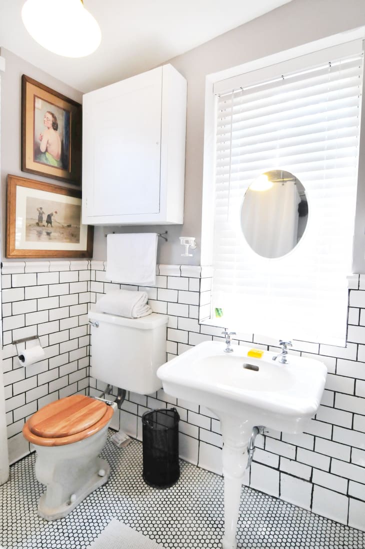 Small bathroom with white subway tiles, wooden toilet seat, pedestal sink, and framed art on the wall.