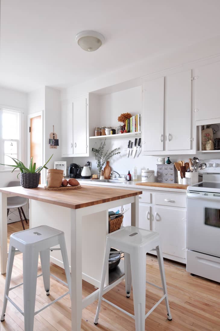 White kitchen with wooden countertops, island with stools, potted plant, and shelves with books and jars.