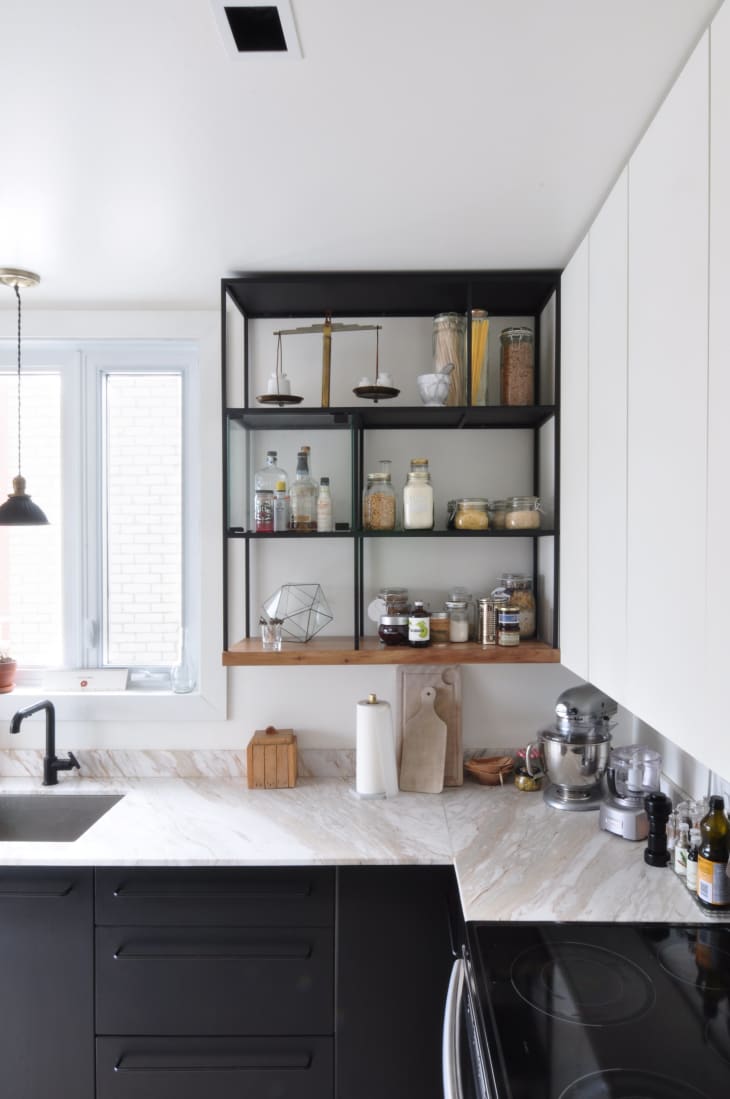 Modern kitchen with black cabinets, marble countertops, open shelves with jars, a mixer, and a window with natural light.
