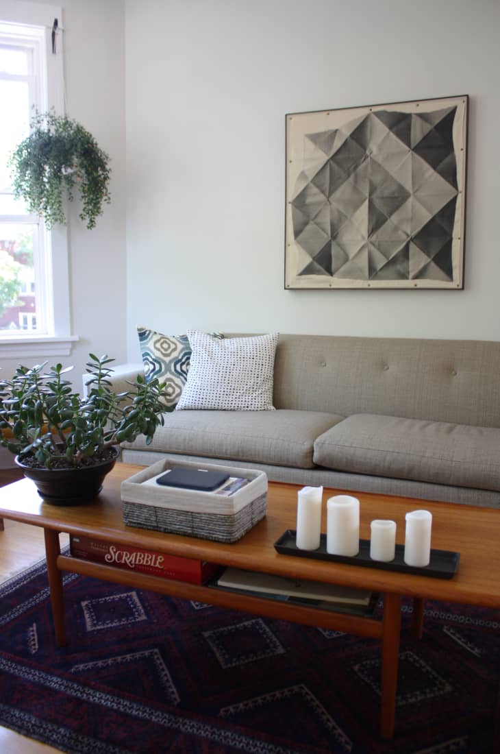 Living room with a gray sofa, geometric wall art, hanging plant, and a wooden coffee table with candles and a Scrabble game.