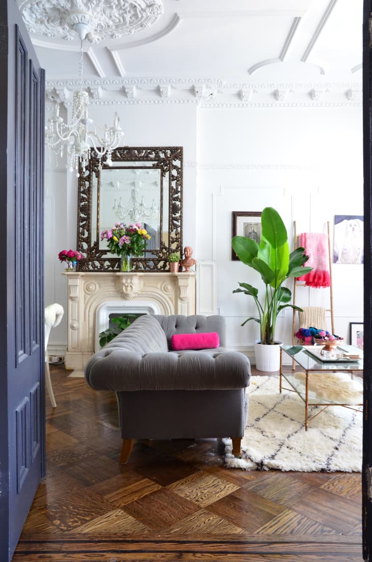 Elegant living room with gray sofa, ornate mirror, chandelier, and vibrant plants on a patterned rug.