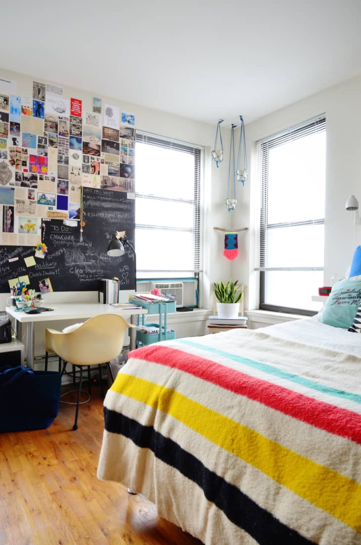 Bedroom with colorful striped bedspread, collage wall, chalkboard, desk, chair, and potted plant by the window.