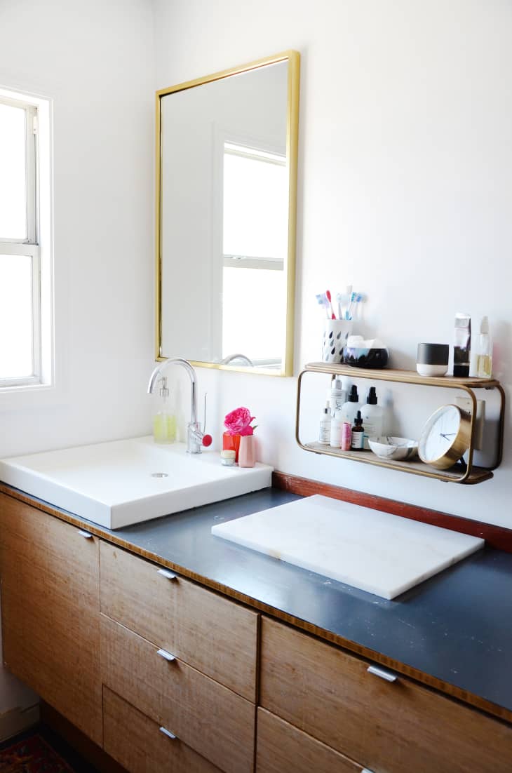 Bathroom vanity with wooden cabinets, large mirror, white sink, and shelf holding toiletries and a clock.