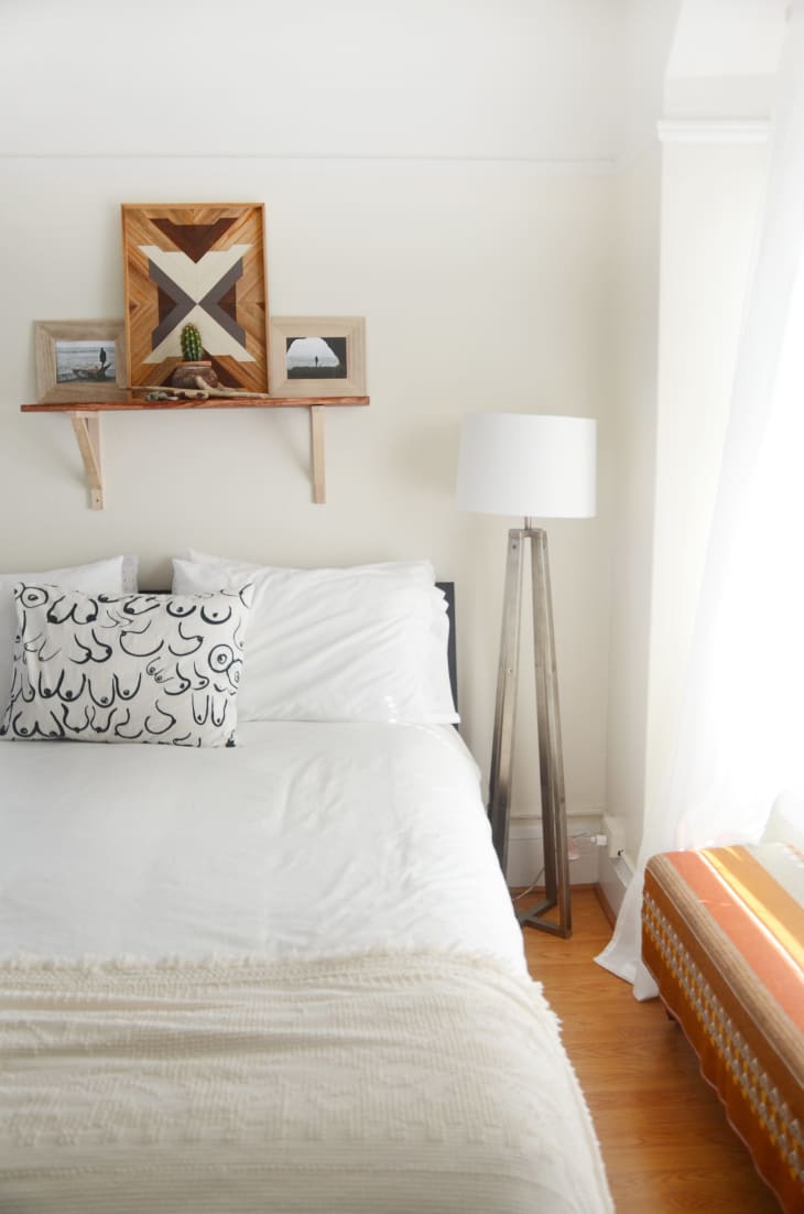 Bedroom with white bedding, abstract pillow, wooden shelf with decor, and a tall floor lamp.