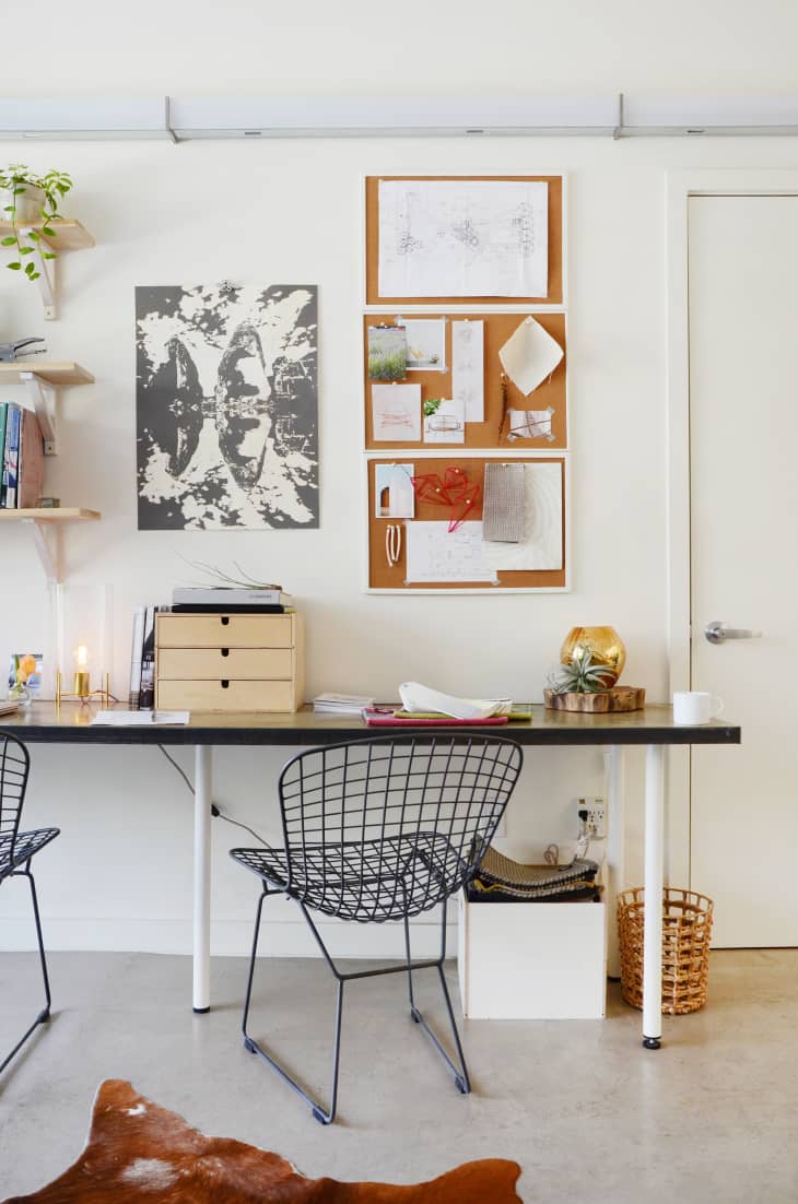 Home office with wire chairs, a long desk, corkboard with notes, abstract art, and a small plant on a shelf.