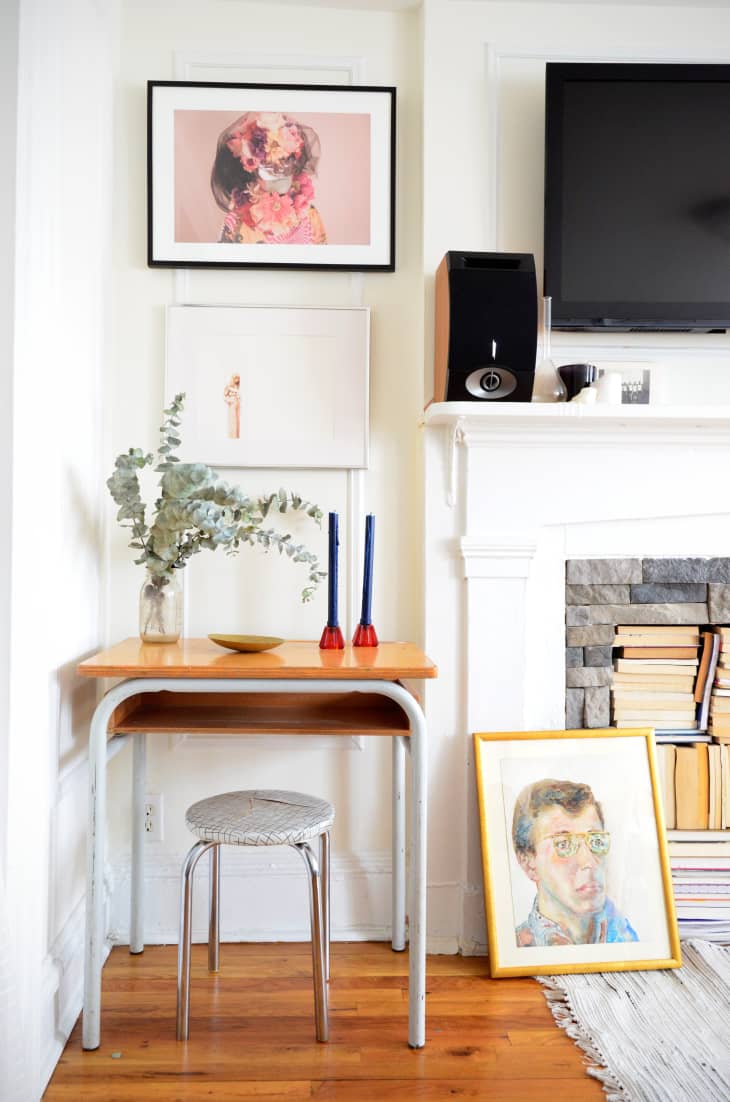 Small desk with eucalyptus vase, blue candles, and framed art near a fireplace with stacked books and a TV above.