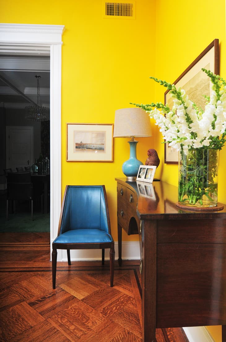 Yellow hallway with a blue chair, wooden sideboard, lamp, framed art, and white flowers in a vase.