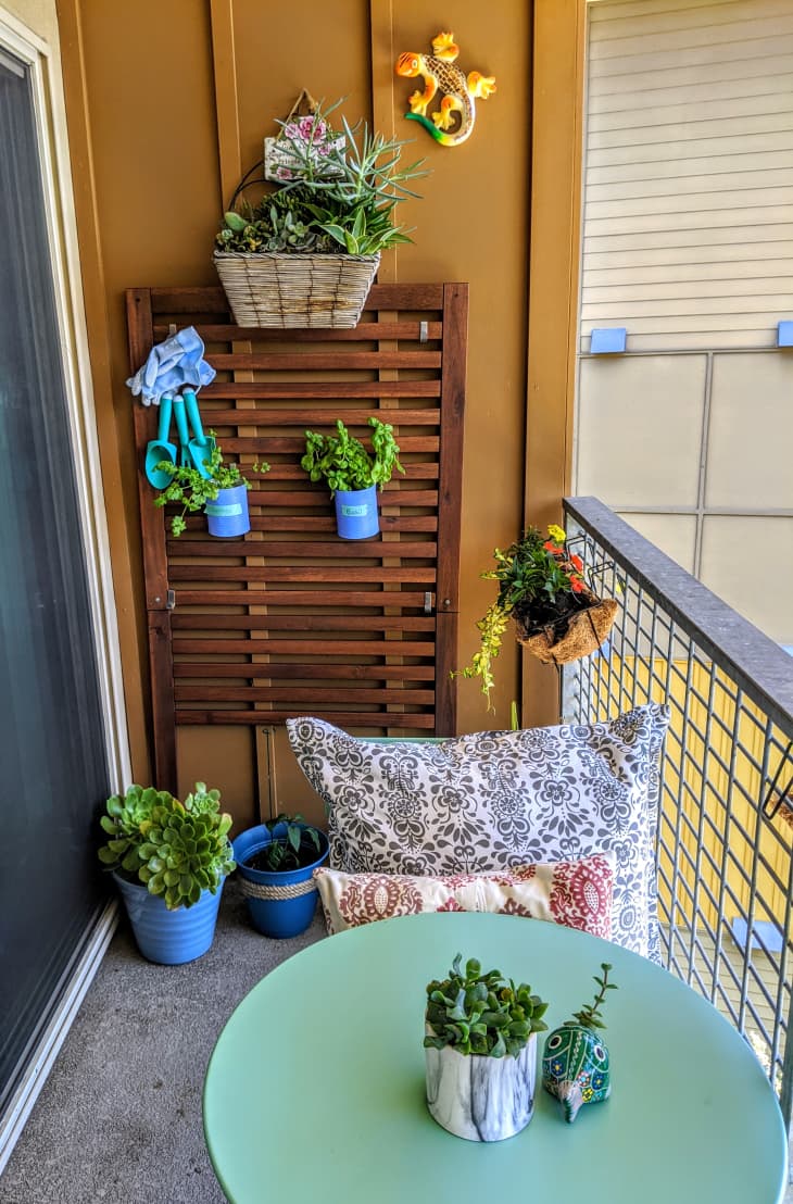 Small balcony with potted plants, a green table, patterned cushions, and a decorative wall gecko.