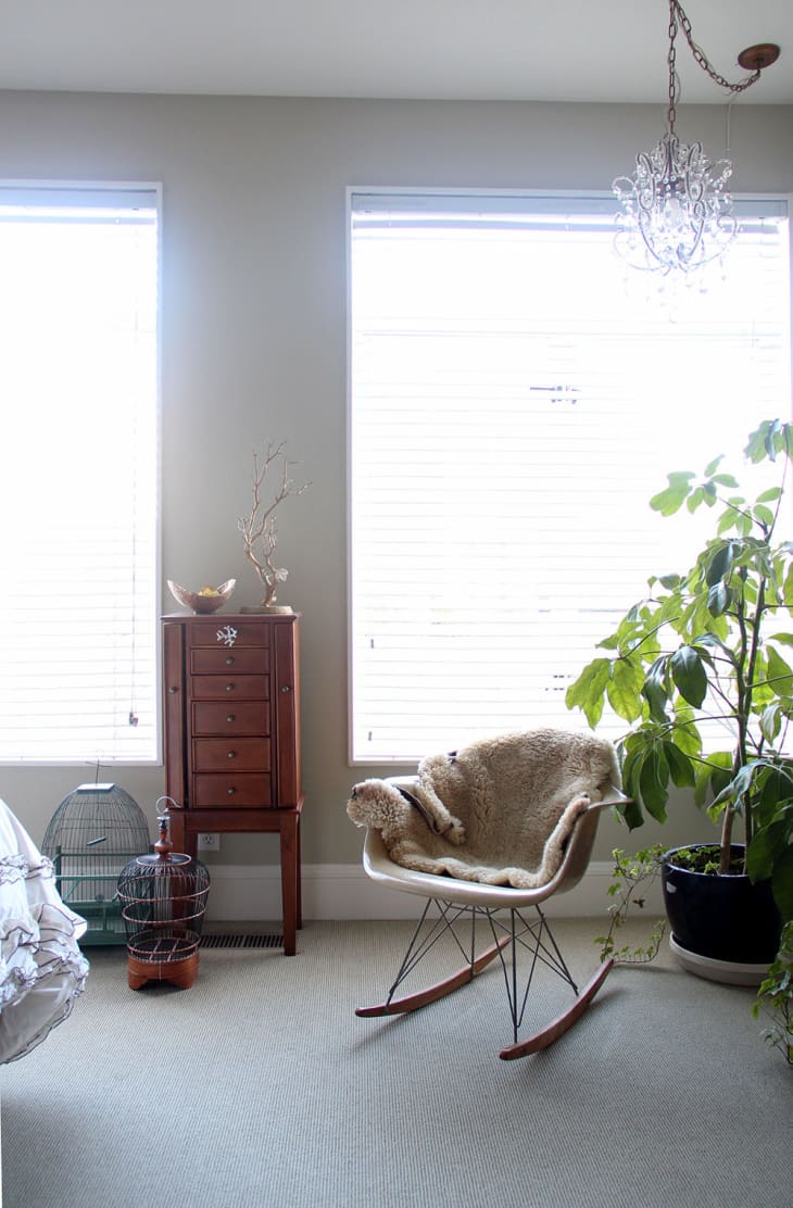 Rocking chair with fur throw, wooden cabinet, birdcage, and large potted plant in a bright room with chandelier.